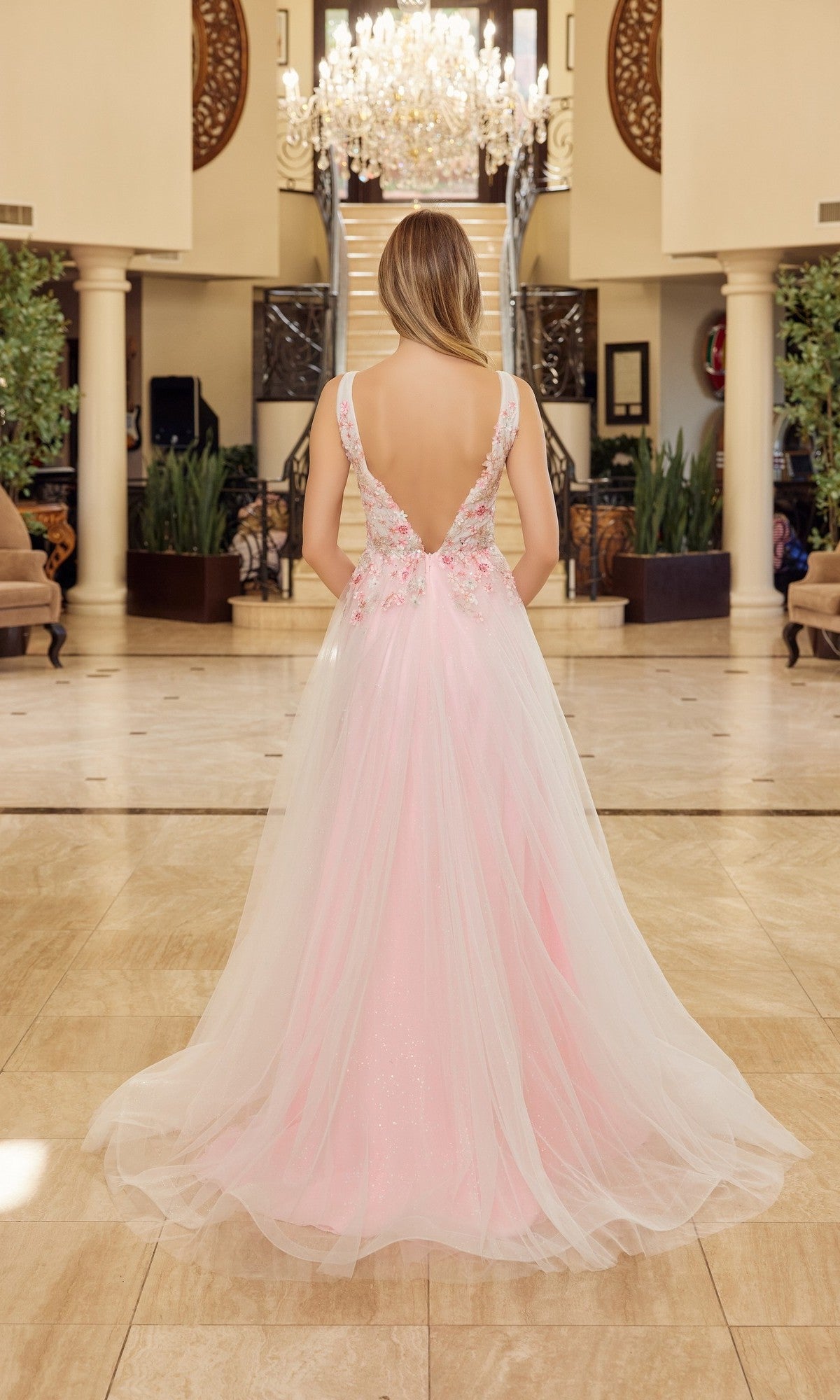 A woman wearing the Nox Anabel Y1799 long prom dress—a pink, floor-length, backless gown with floral details—stands in an elegant room with a marble floor and chandelier.